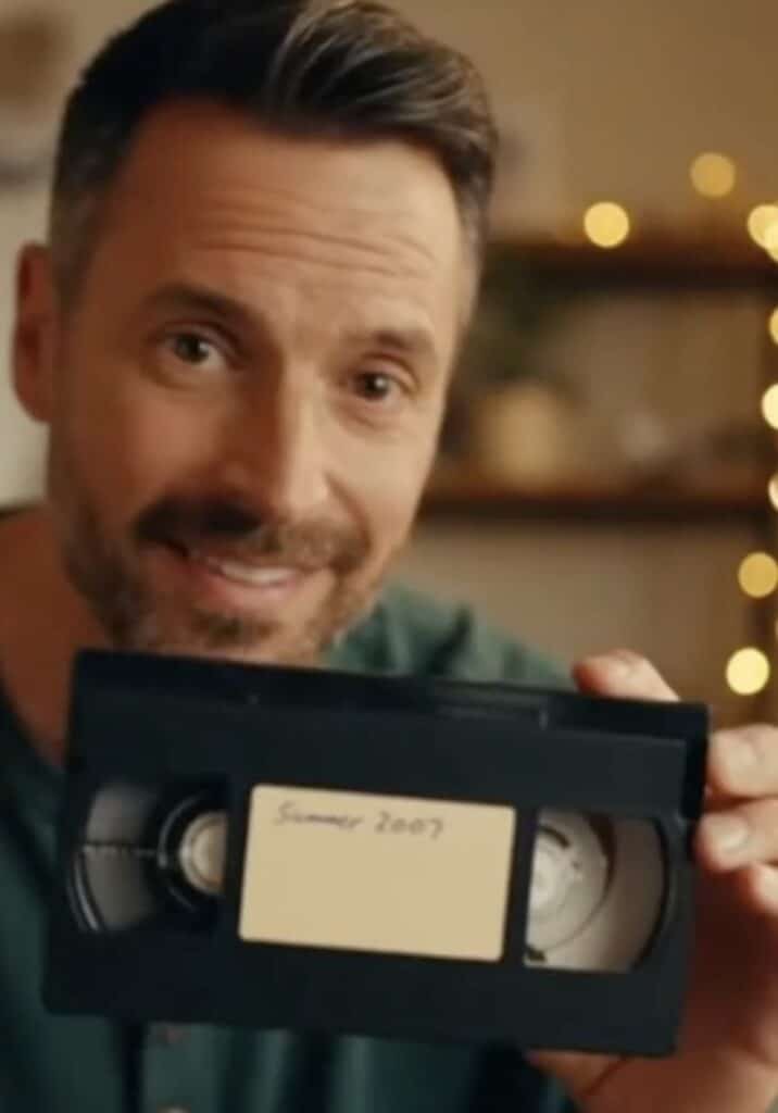 A smiling man holds up a black VHS tape labeled "Summer 2007" in front of a blurred background with string lights. The man has short brown hair, a beard, and is wearing a green shirt.